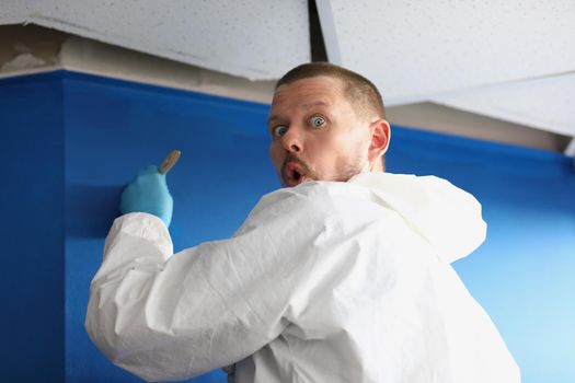 Portrait of male worker caught by surprise painting wall in blue colour. Carpenter in uniform use brush for applying paint. House design project concept