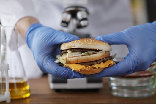 Close-up of male chemist holding tasty burger in hand with blue protective glove. Lab experiment on fastfood product. Health study and testing food concept