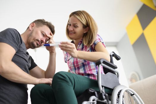 Low angle of happy female sit in wheelchair and show to husband positive pregnancy test. Upset man not happy, confused emotion. Disability, family concept