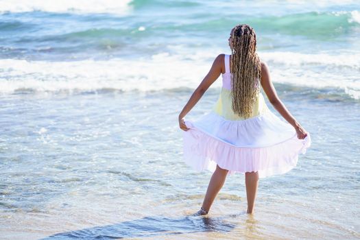 Black girl walking along the shore of the beach wearing a beautiful long dress. Young girl enjoying her vacations.