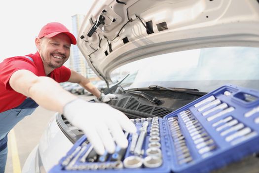Portrait of automotive technician smiling and take instrument from open kit. Open car hood and need to fix problem of automobile. Car maintenance concept