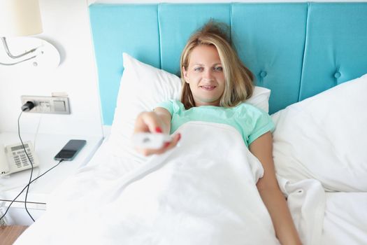Portrait of happy woman laying in bed and use remote control to change channel on tv. Rest in hotel room, stylish interior, summer resort. Chill concept
