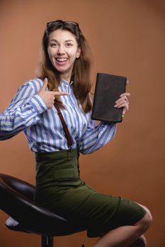 Attractive young Caucasian woman in office style sits on a chair laughing and points to her diary. Studio portrait.