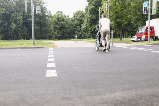 Low angle of man drive woman on wheelchair across street at pedestrian crossing. Caregiver walk with patient. Disabled people, accident, recovery concept