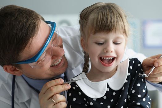 Portrait of man pediatrician check little girls health condition using special tool for mouth. Qualified family doctor provide diagnostic. Medicine concept
