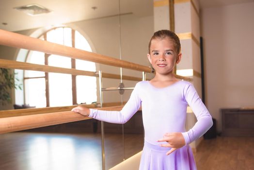 a little smiling girl in pointe shoes does stretching in ballet class near a frame and a big mirror