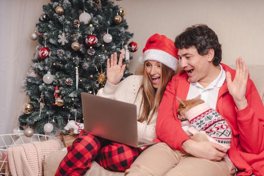 Portrait of lovely couple husband and wife with laptop in red Santa hat having a video call chat with puppy dog in sweater, enjoy Christmas time at home. Online greeting