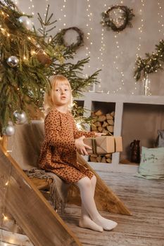 A little blonde girl is sitting on a wooden staircase in a Scandinavian interior decorated in a New Year's style. A child holds a Christmas gift in a craft package.