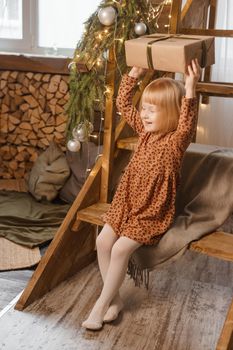 A little blonde girl is sitting on a wooden staircase in a Scandinavian interior decorated in a New Year's style. A child holds a Christmas gift in a craft package.