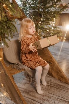 A little blonde girl is sitting on a wooden staircase in a Scandinavian interior decorated in a New Year's style. A child holds a Christmas gift in a craft package.