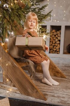 A little blonde girl is sitting on a wooden staircase in a Scandinavian interior decorated in a New Year's style. A child holds a Christmas gift in a craft package.