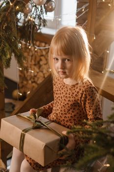 A little blonde girl is sitting on a wooden staircase in a Scandinavian interior decorated in a New Year's style. A child holds a Christmas gift in a craft package.