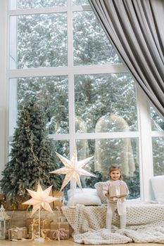 Happy beautiful girl take present in front of big window. Christmas time. It's snow outside. Christmas tree.