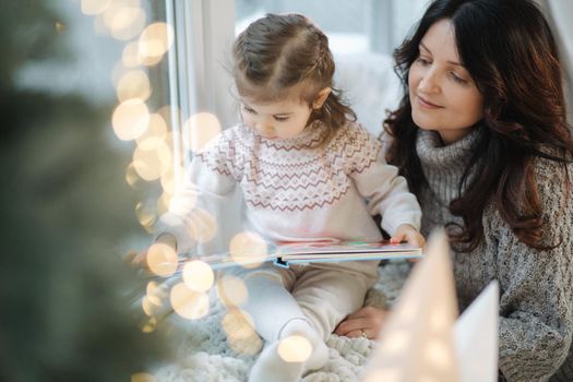 Young grandmother play and read book with her adorable grandaugher nea fir tree. Christmas tree