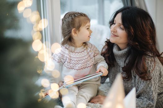 Young grandmother play and read book with her adorable grandaugher nea fir tree. Christmas tree