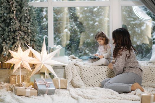 Young grandmother play and read book with her adorable grandaugher nea fir tree. Christmas tree