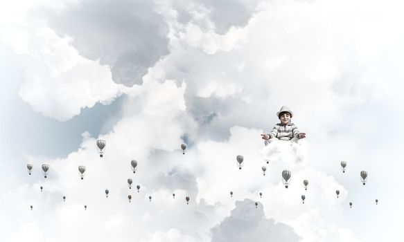 Young little boy keeping eyes closed and looking concentrated while meditating among flying balloons in the air with cloudy skyscape on background.