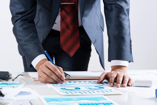 Businessman standing near office desk and analyzing financial charts. Successful corporate manager in business suit and tie. Close-up man hand with pen. Entrepreneurship and leadership concept.