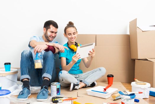 Happy couple sitting on floor and looking at tablet computer screen. Home remodeling and house interior redesign. Painting tools and materials on floor. Young family renovating their home after moving