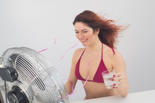 Red-haired smiling woman in a bikini drinks a cold drink and enjoys the blowing wind from an electric fan on a white background. Climate control on a hot summer day.