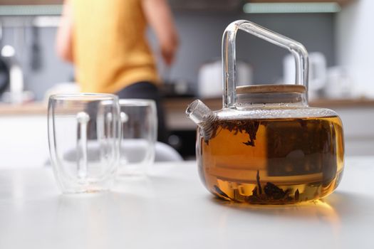 A woman made tea in a glass teapot, close-up, blurry. Glassware in the kitchen, coziness, hot herbal drink