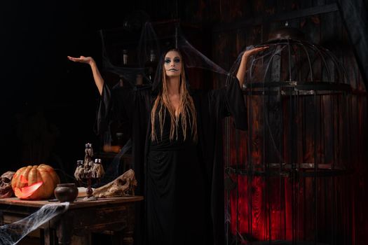 Halloween concept. Witch dressed black hood with dreadlocks standing dark dungeon room rise hand for conjuring magic spell. Female necromancer wizard rise up hands skull, cage, spider web