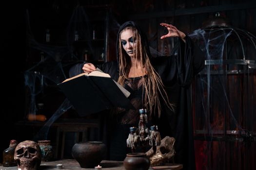 Halloween concept. Witch dressed black hood standing dark dungeon room use magic book for conjuring magic spell. Female necromancer wizard gothic interior with skull, cage, spider web