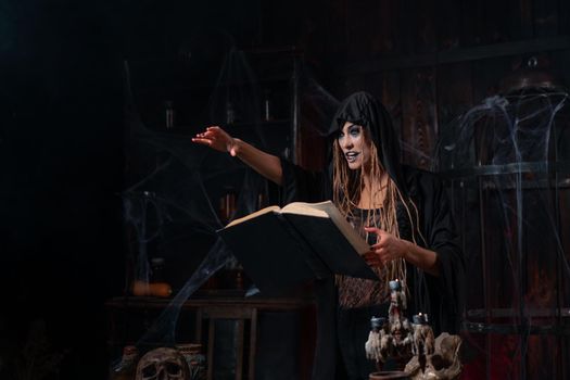 Halloween concept. Witch dressed black hood standing dark dungeon room put her hand forward use magic book for conjuring magic spell. Female necromancer wizard gothic interior Devil inside