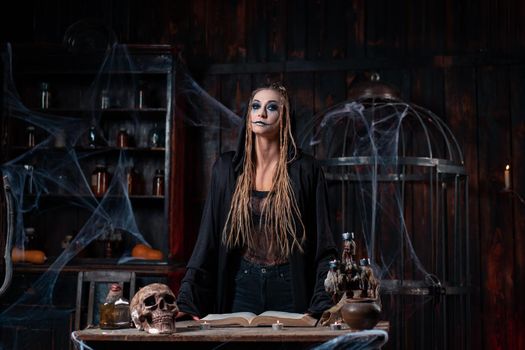 Halloween concept. Witch dressed black hood with dreadlocks standing dark dungeon room use magic book for conjuring magic spell. Female necromancer wizard gothic interior with skull, cage, spider web