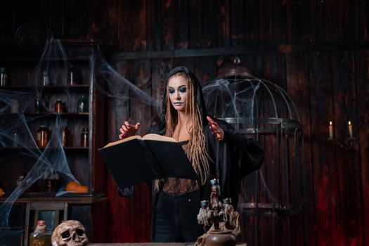 Halloween concept. Witch dressed black hood standing dark dungeon room use levitating magic book conjuring magic spell. Female necromancer wizard gothic interior with skull, cage, spider web