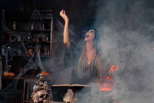 Halloween, witch use magic book and cauldron prepare poison or love potion. Black magic occult female wizard in dark room cage spider web human skull. Necromancer screaming raised hand. voodoo magic