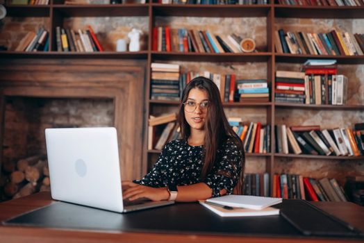 Young beautiful woman in glasses works at the laptop while sitting at the table