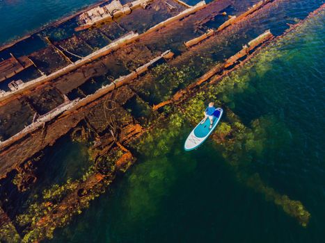 Woman on paddle board, sup next to Abandoned broken shipwreck sticking out of the sea.