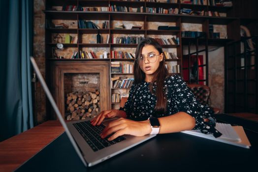 Young beautiful woman in glasses works at the laptop while sitting at the table