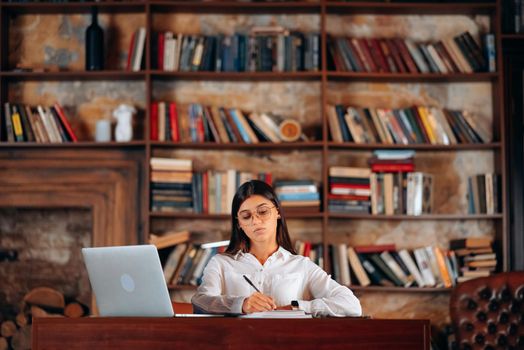 Young beautiful woman in glasses writes in a notebook while sitting at the table