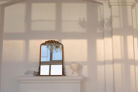Mirror placed on artificial white fireplace near statuette and stack of books against white wall with shadow of window and chandelier on it