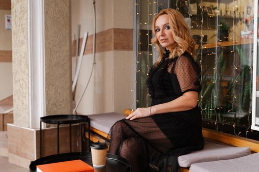 Young charming blonde with a cute smile and makeup while relaxing in a cafe. She is holding a cup of coffee in her hands. She is dressed in a black dress with transparent sleeves