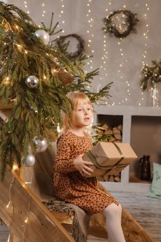 A little blonde girl is sitting on a wooden staircase in a Scandinavian interior decorated in a New Year's style. A child holds a Christmas gift in a craft package.