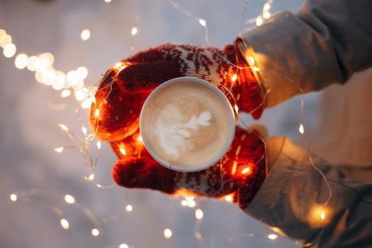 Holds coffee in red mittens on a snowy background with garlands on top. High quality photo