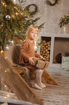 A little blonde girl is sitting on a wooden staircase in a Scandinavian interior decorated in a New Year's style. A child holds a Christmas gift in a craft package.