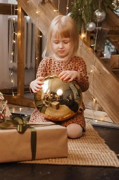 A little blonde girl is sitting next to a wooden staircase in a Scandinavian interior decorated in a New Year's style. A child holds a golden Christmas ball.