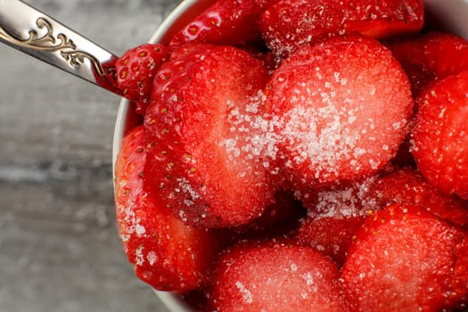 Detailed top view on small bowl with fresh cut strawberries sprinkled with white crystal sugar, silver spoon suggesting they're ready to eat.