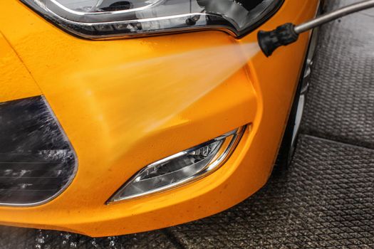 Detail on yellow car front bumper and light being washed with jet water spray in carwash.