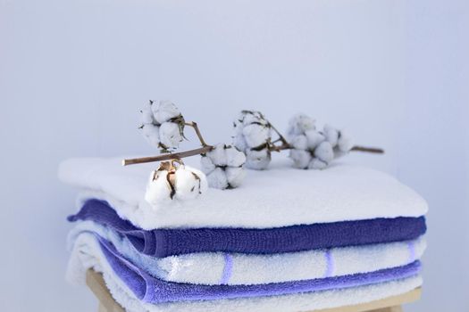 stack of clean bath towels colorful cotton Terry textiles with a sprig of cotton folded on a wooden chair against a white wall nap concept close-up country style. Demonstrating the colors of 2022 - Very Peri