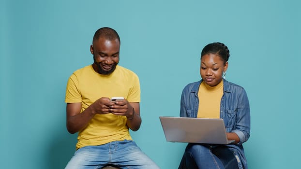 Man and woman using smartphone and laptop in front of camera while they sit together. Modern couple working with mobile phone and computer on online internet to have fun over blue background.