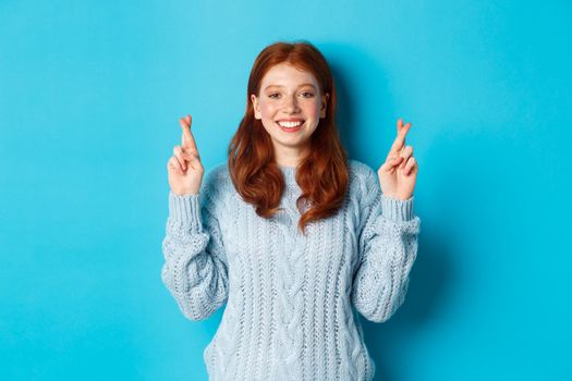 Hopeful redhead girl making a wish, cross fingers for good luck, smiling and anticipating good news or positive result, standing against blue background.