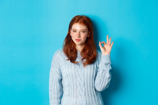Confident redhead girl assuring you, showing okay sign and smiling, saying yes, approve and agree, standing over blue background.