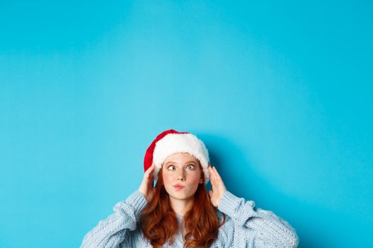 Winter holidays and Christmas eve concept. Head of funny redhead girl in santa hat, appear from bottom and squinting, making silly faces, standing near copy space on blue background.