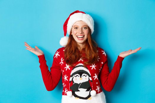 Happy holidays and Christmas concept. Cheerful redhead girl in Santa hat and xmas sweater, raising hands on copy spaces, holding something on blue background.