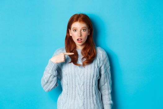 Confused redhead girl pointing at herself, being chosen, standing in sweater against blue background.